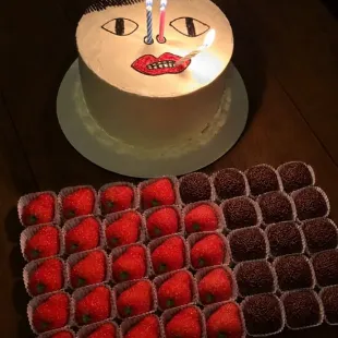 Bolo com rosto desenhado e velas, ao lado de uma bandeja de morangos e brigadeiros